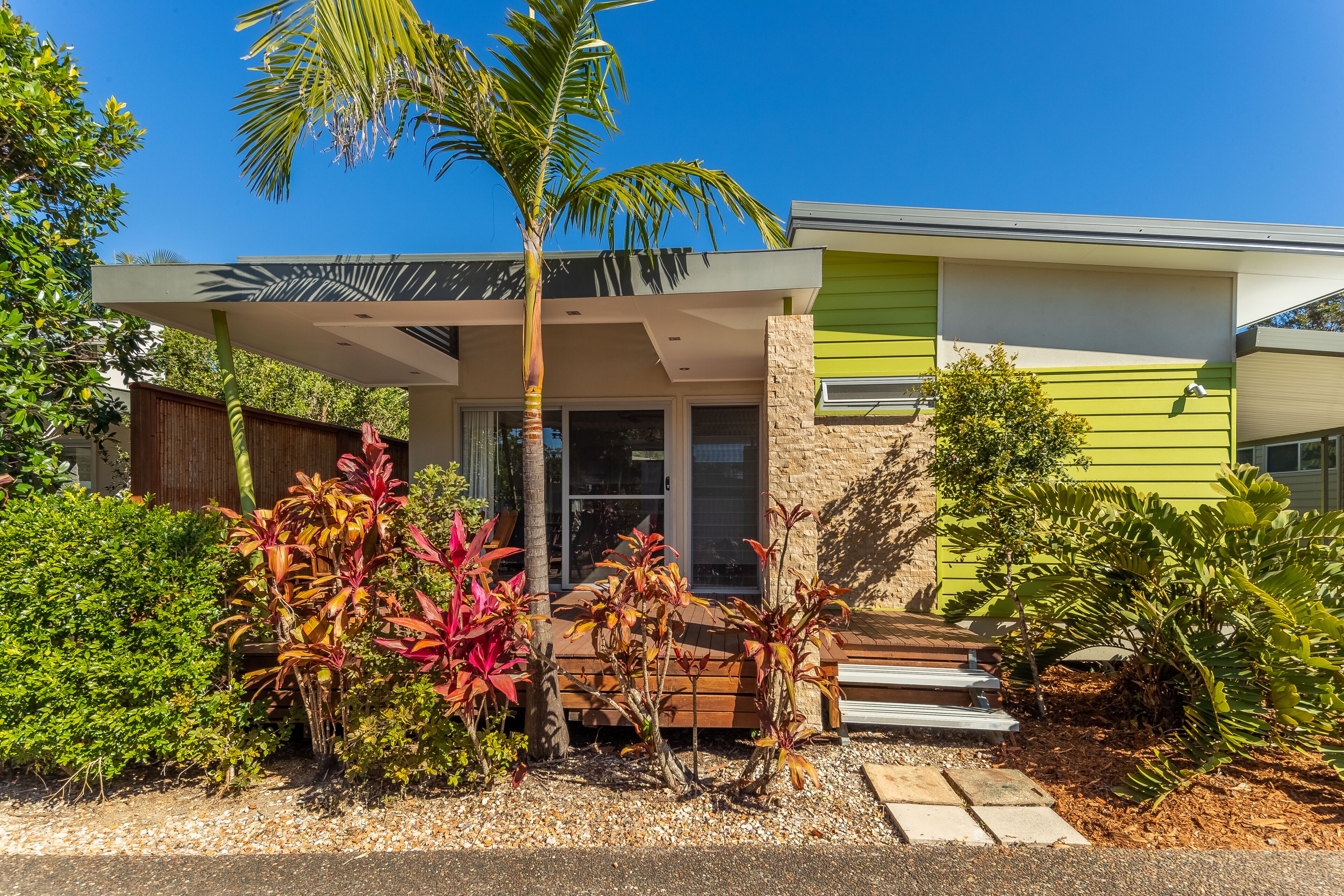 A modern house with a covered porch, a stone wall, and a variety of tropical plants.