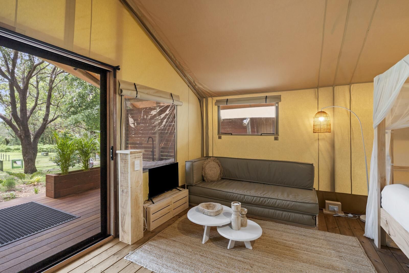 A cozy room with a wooden floor, a sofa, a small table, a TV, and a window overlooking greenery.
