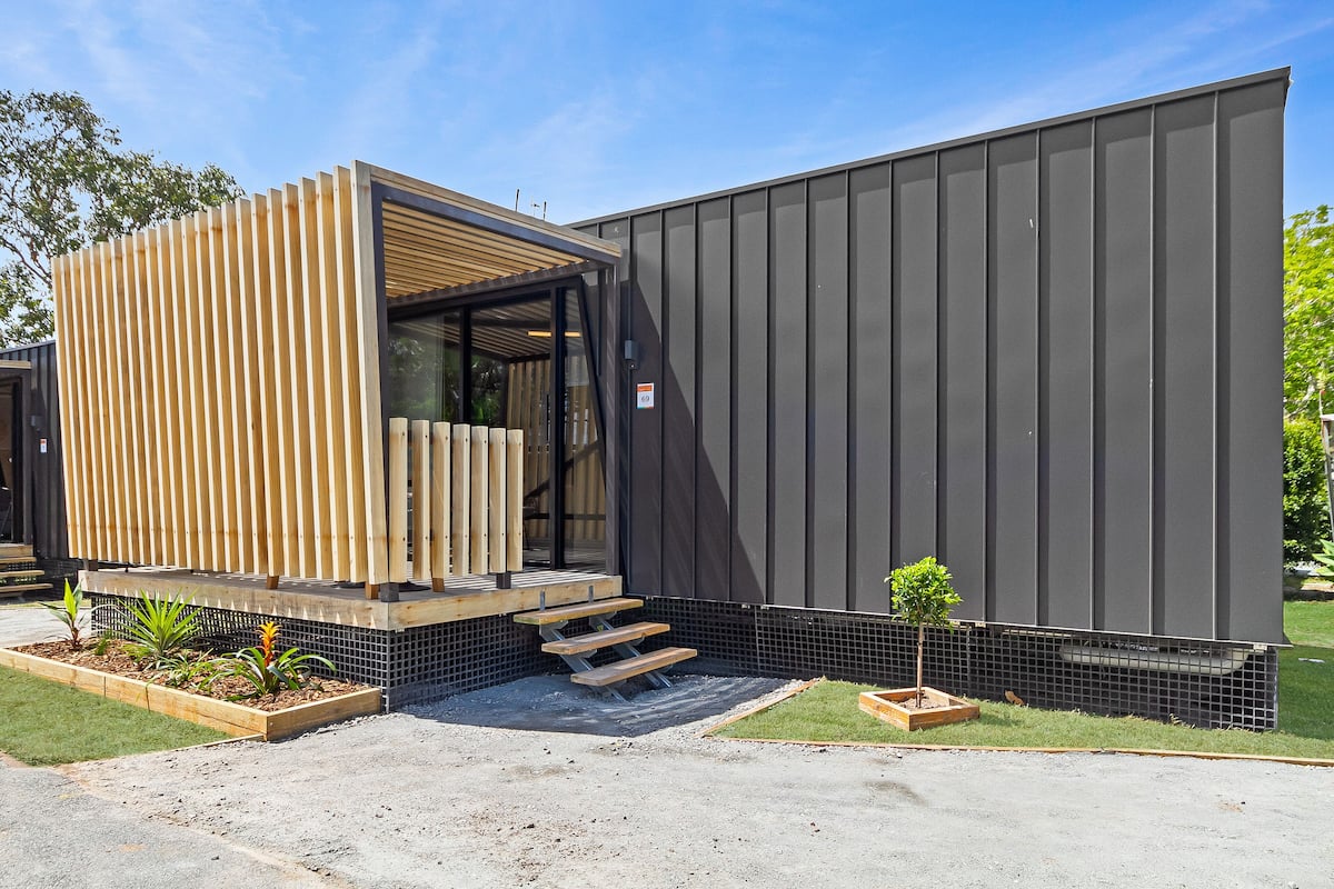 Ein modernes Gebäude mit einer Holz-Jalousiefassade, einer kleinen Außen-Sitzgelegenheit und einer metallartigen Containerstruktur.