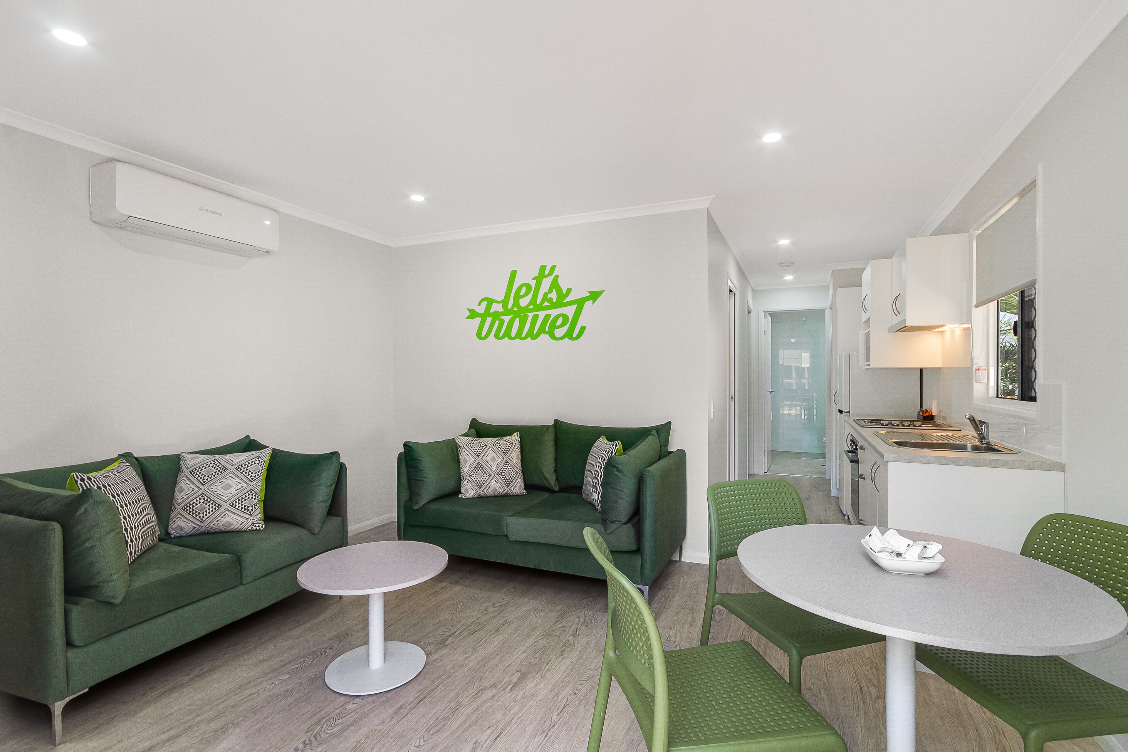 Un salon moderne avec deux canapés verts, une table ronde blanche et un coin cuisine comprenant un évier et des placards.