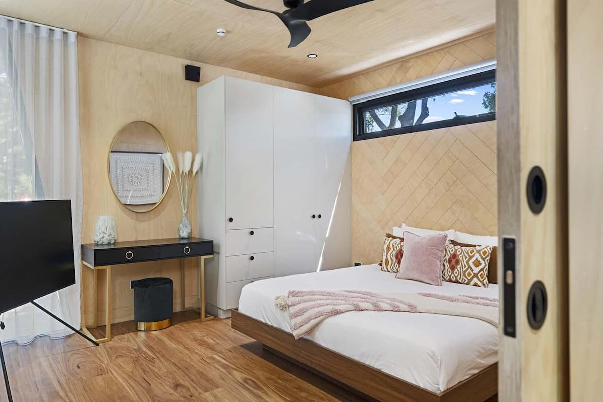 Ein modernes Schlafzimmer mit Holzboden, einem Bett mit gemusterten Kissen, einem Nachttisch mit einer Vase und einem Fernseher.