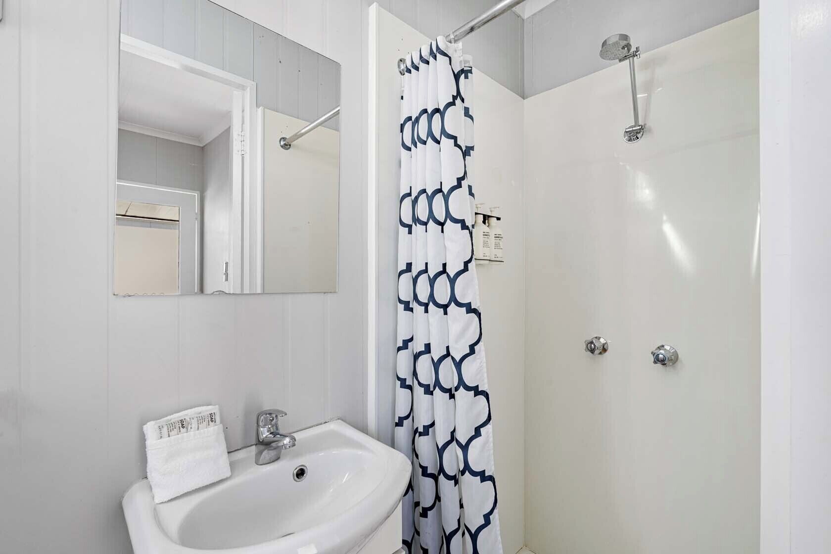 Un baño con un lavamanos blanco, un espejo, una ducha con cortina estampada y una puerta con pomo.