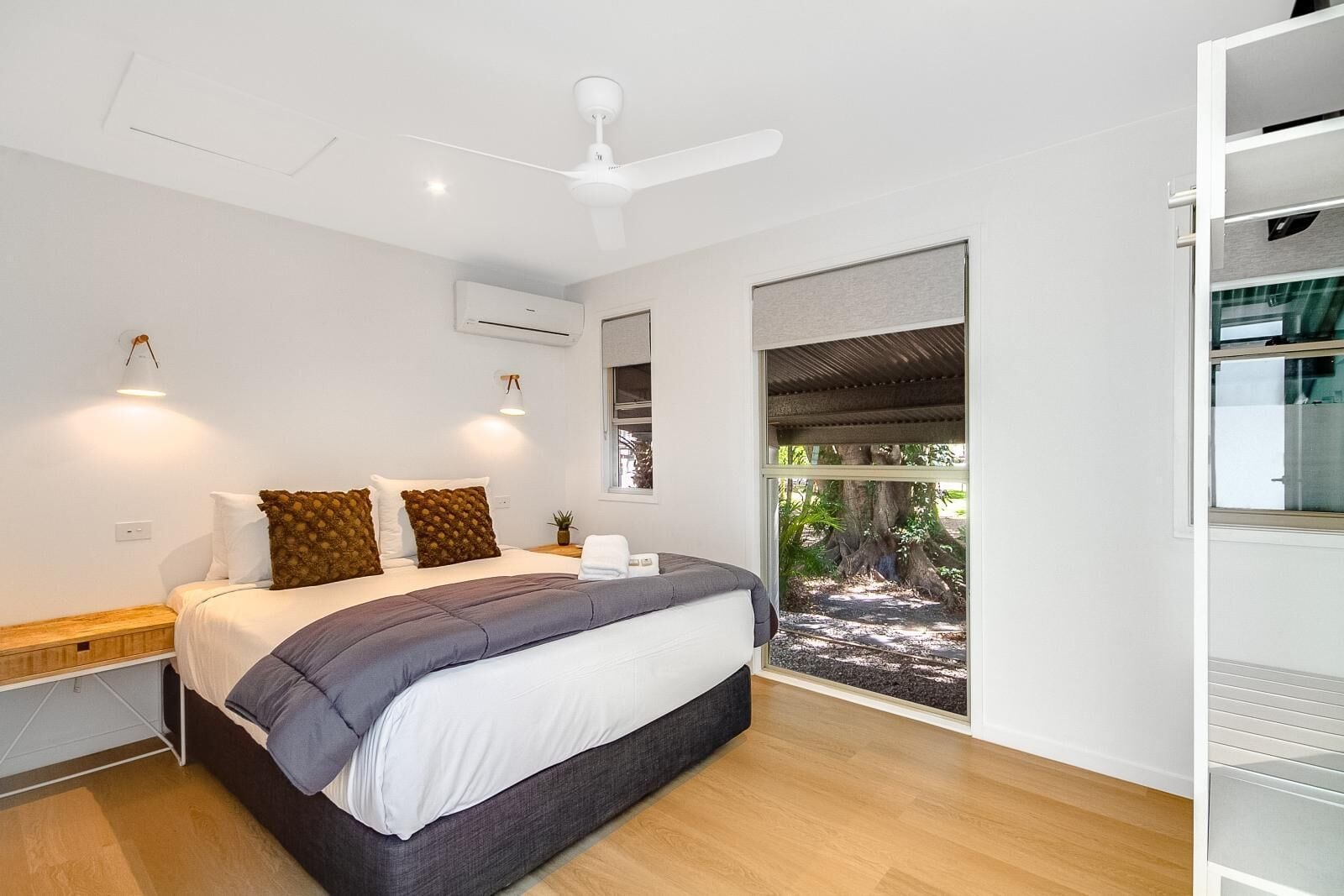 Un dormitorio moderno con una cama grande, un ventilador de techo, un equipo de aire acondicionado y vista a un árbol por la ventana.