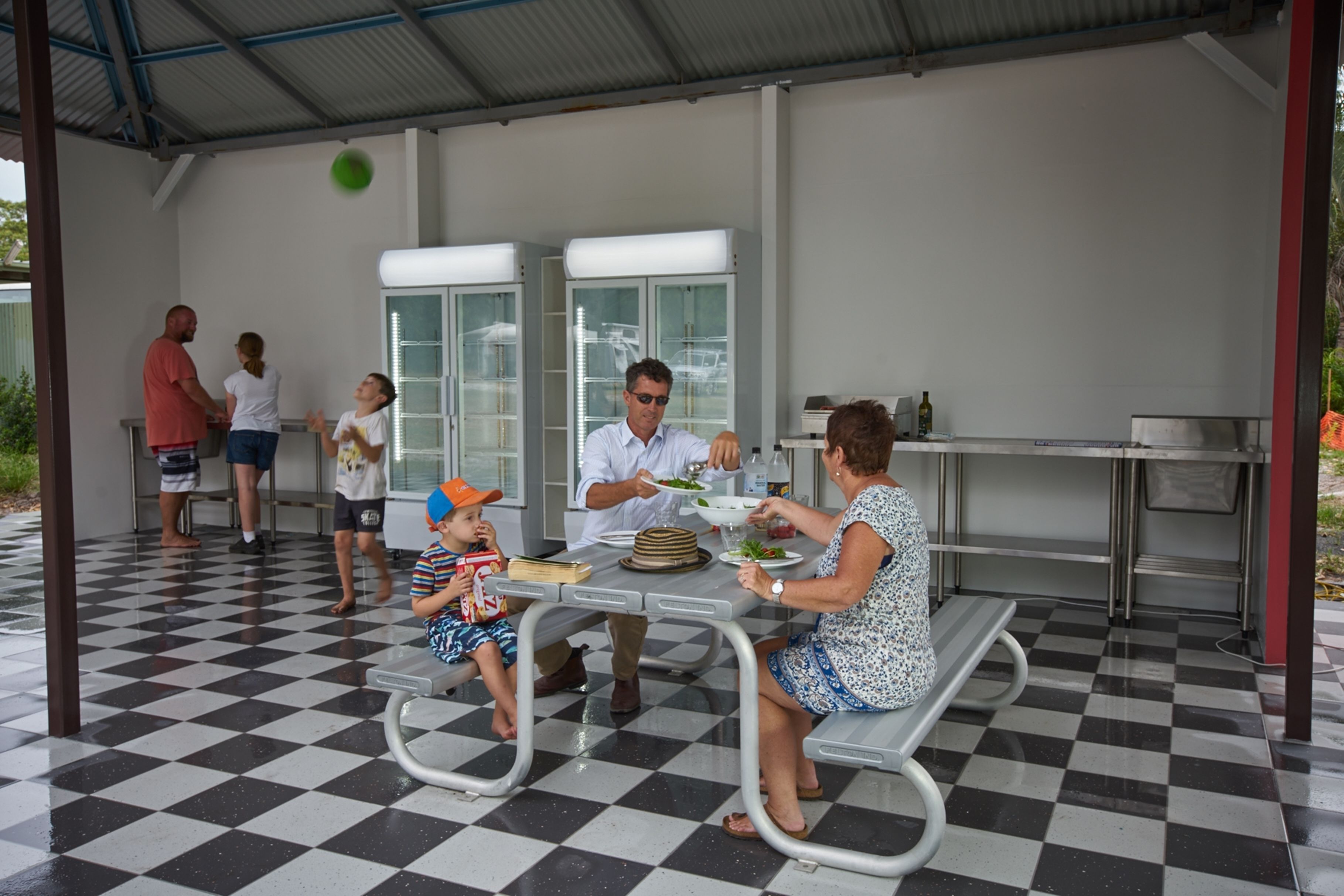 A family dining at an outdoor table with a checkered floor, under a covered area with a metal roof.