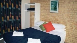 A hotel room with a brick wall, a bed with white linens and two pillows, a white wardrobe, and a framed picture on the wall.