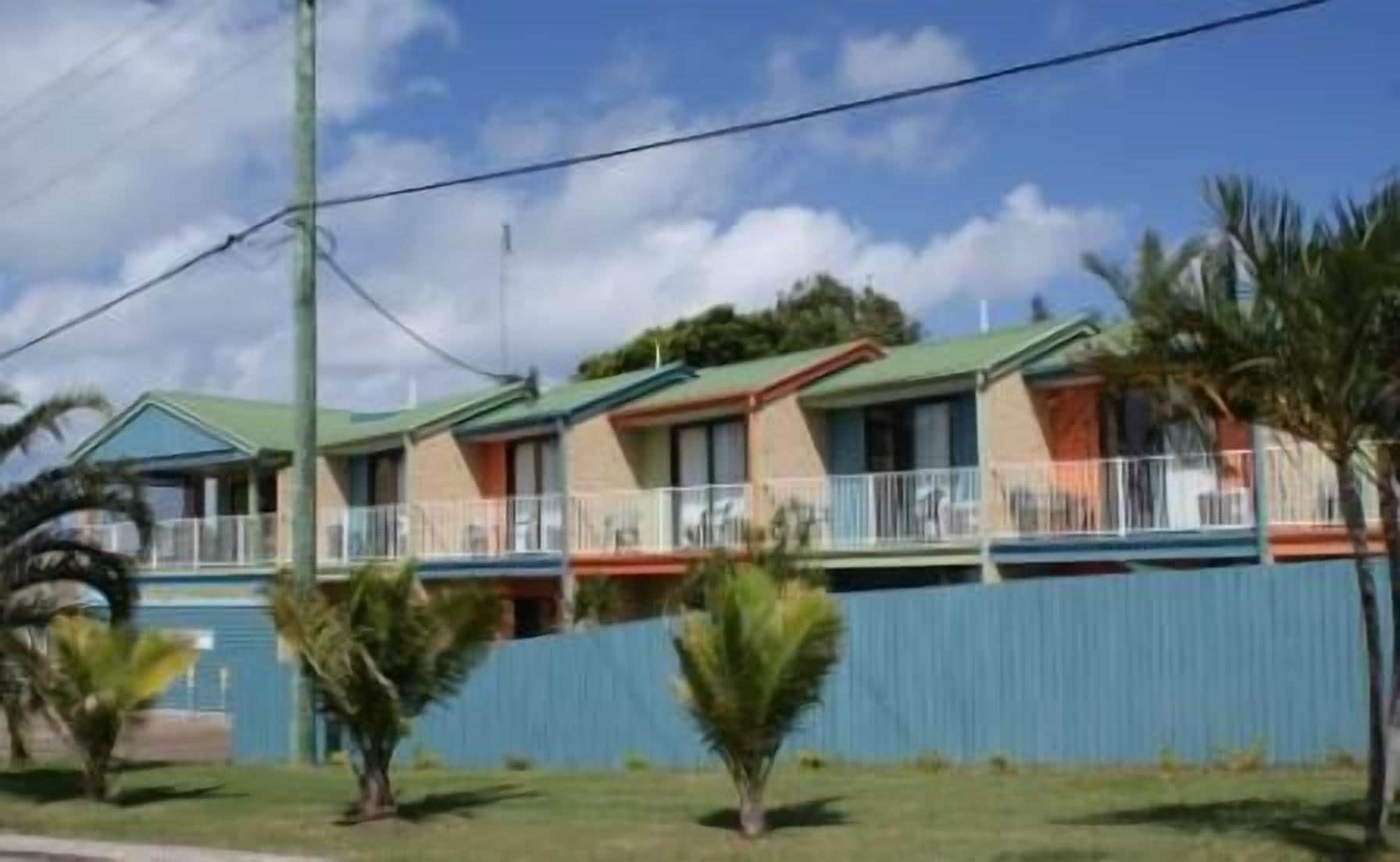 A row of two-story motel buildings with balconies, a blue fence, and palm trees in the foreground.