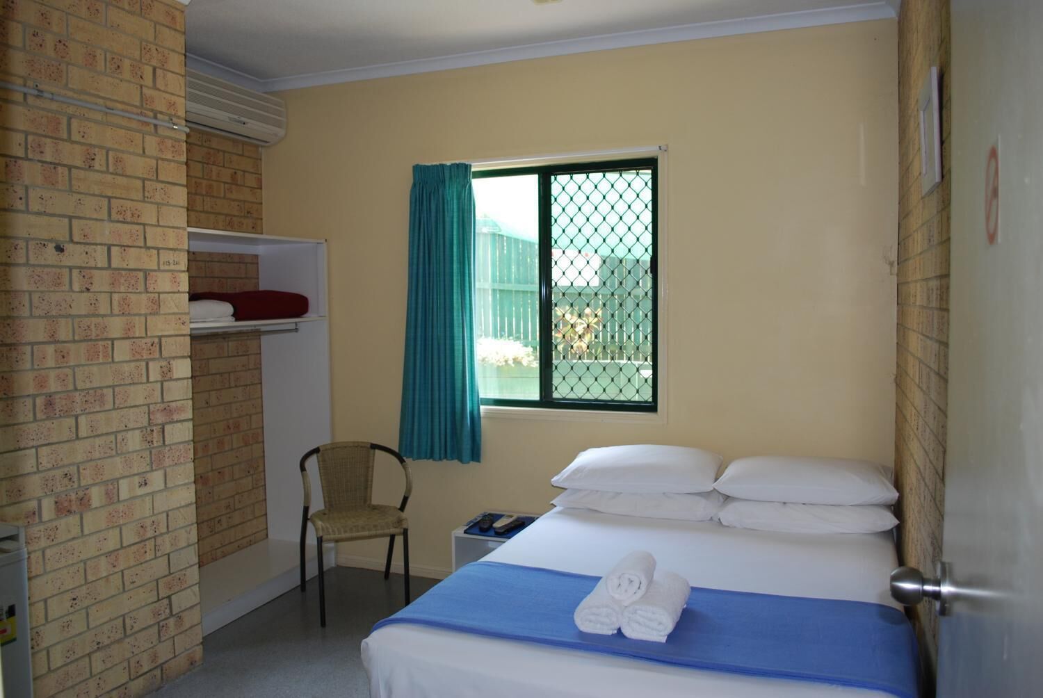 Habitació d'hotel amb un llit, una cadira, una finestra amb cortina i una estant amb un objecte vermell.