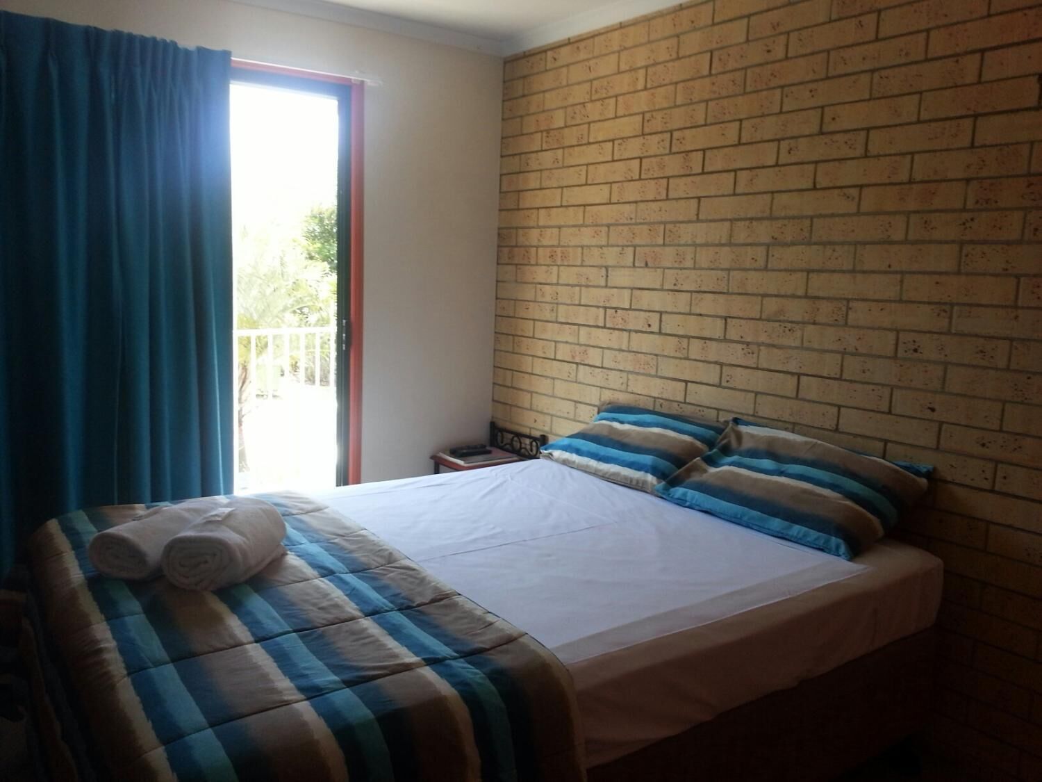 Een slaapkamer met een bed, een raam met gordijnen, een bakstenen muur en een klein tafeltje met een boek.