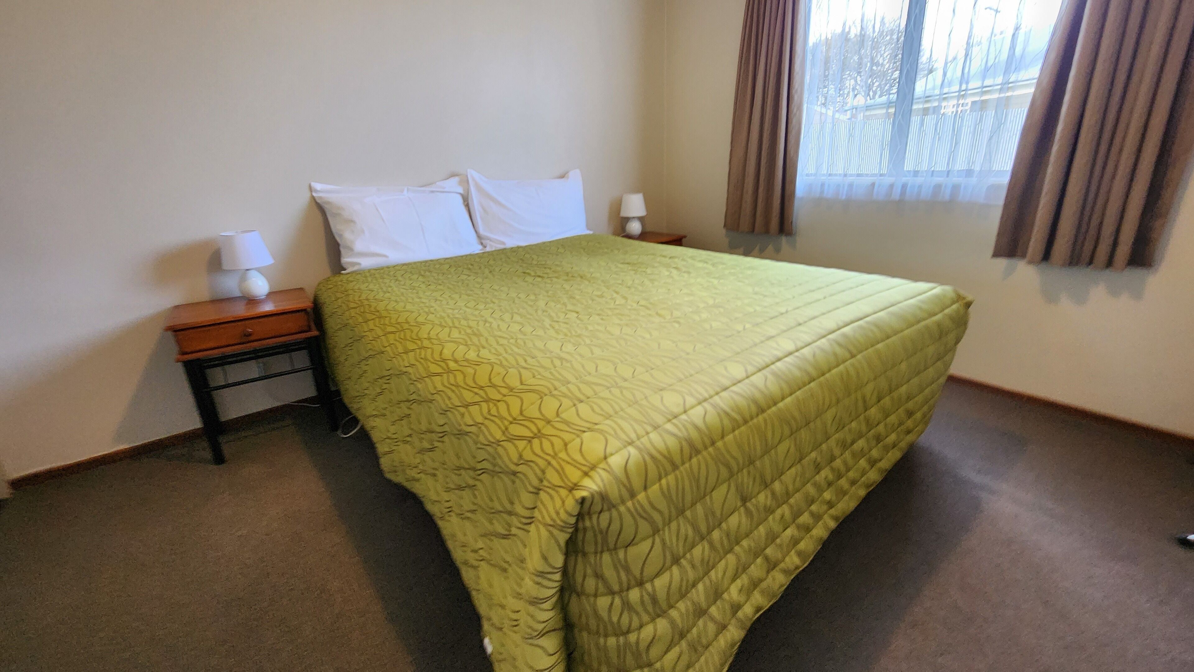 A neatly made bed with a green quilt, two bedside tables with lamps, and a window with curtains.