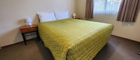 A neatly made bed with a green quilt, two bedside tables with lamps, and a window with curtains.