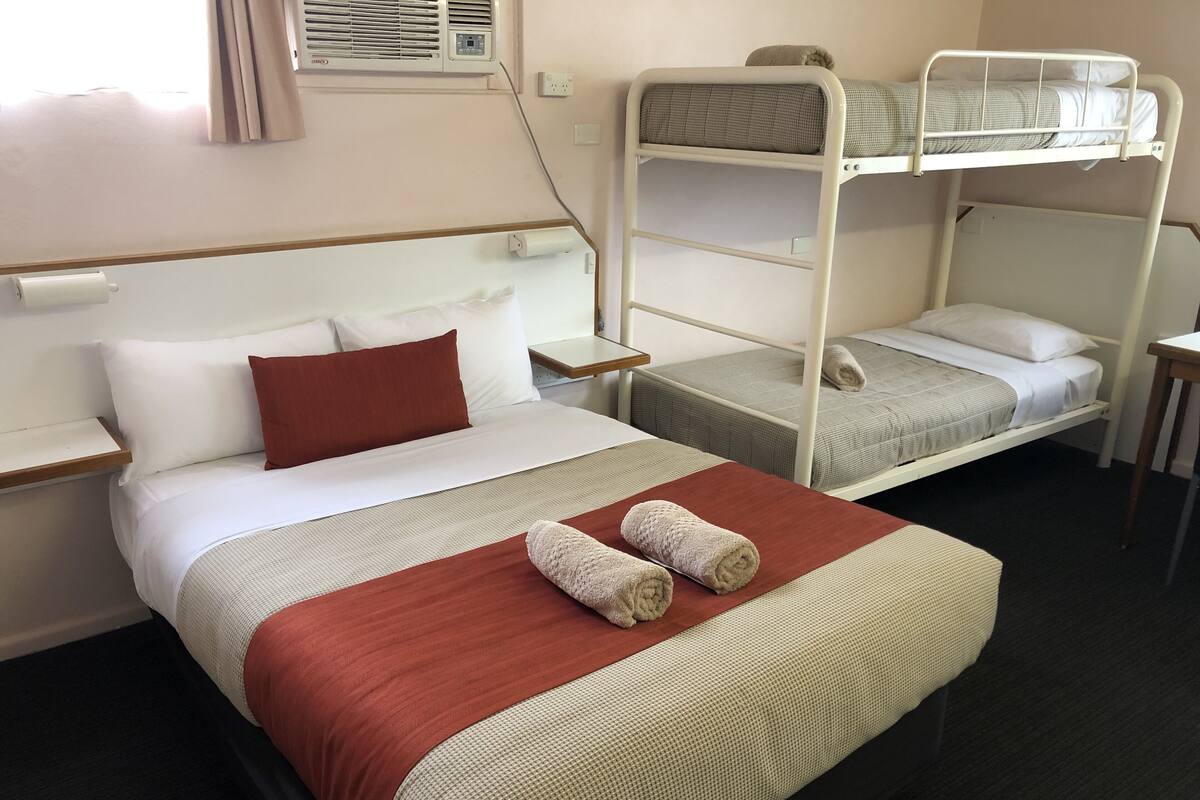 A hotel room with a single bed, a bunk bed, a bedside table, and a wall-mounted air conditioning unit.