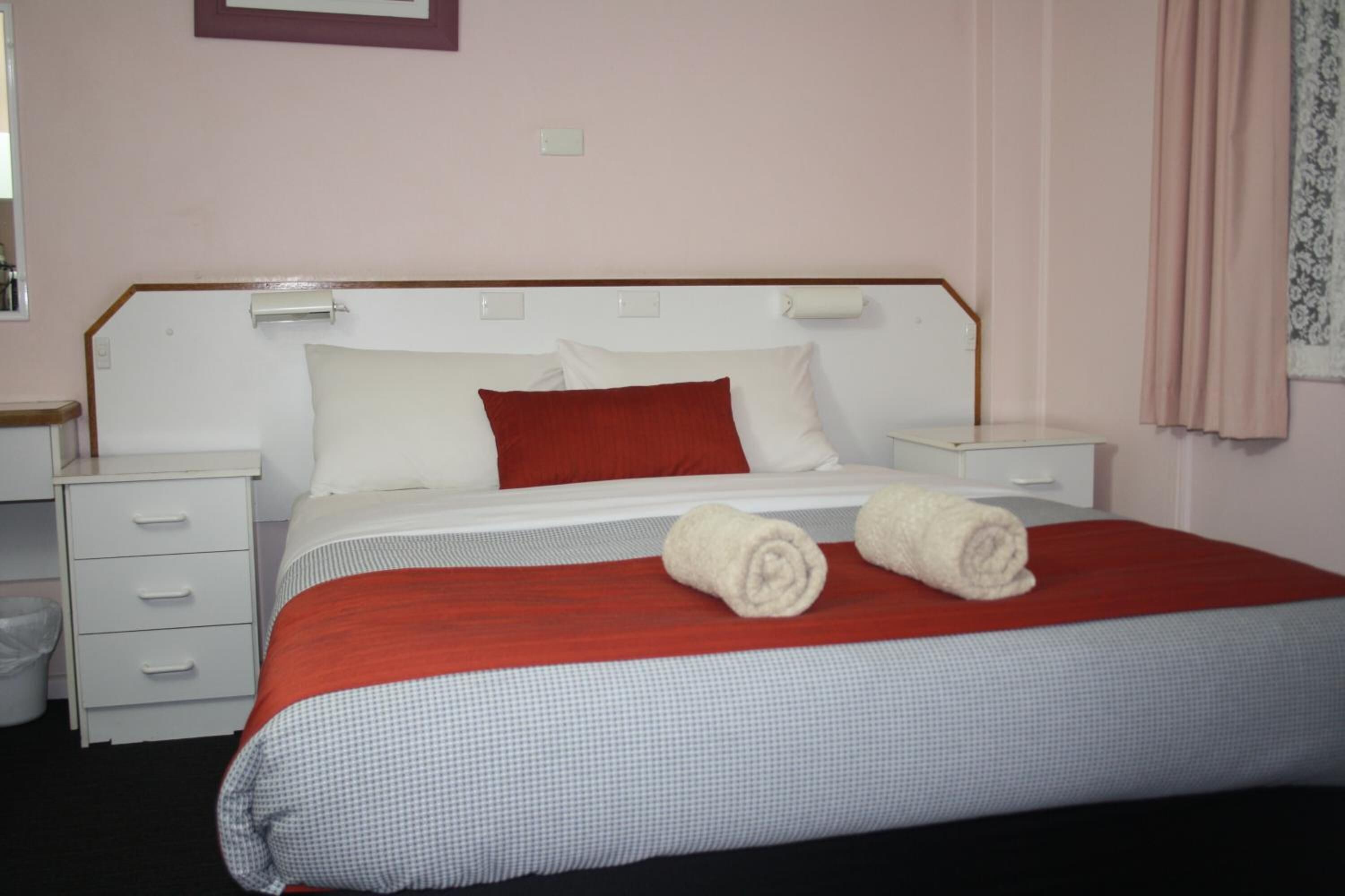 A neatly made bed with a red and white checkered bedspread, two rolled towels, and a white headboard.