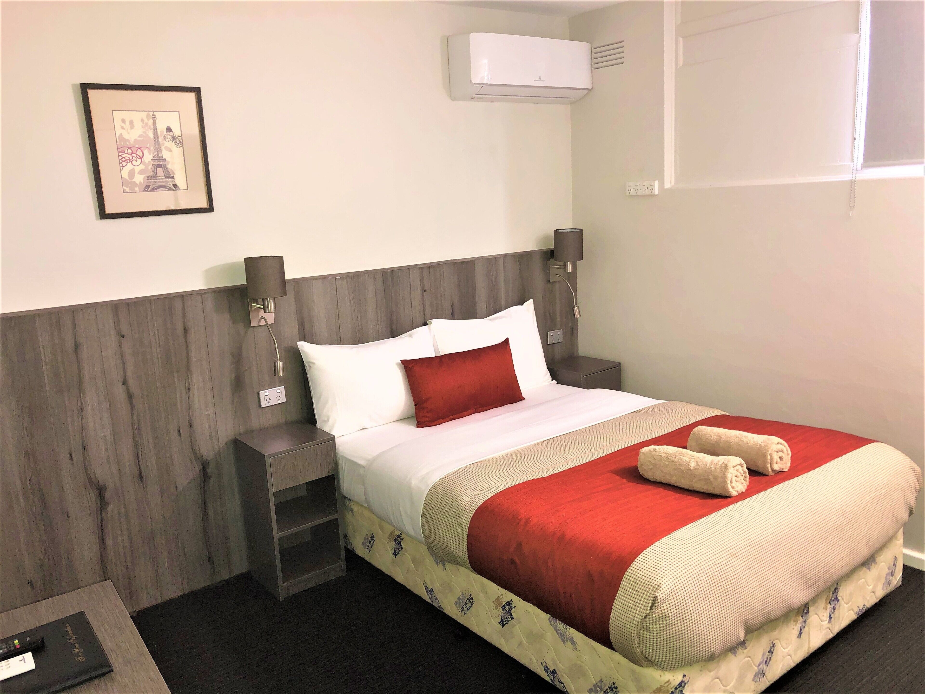 A neatly made bed with a red pillow, a white pillow, and a red blanket. There is a small nightstand with a lamp and a framed picture on the wall.