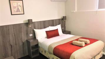 A neatly made bed with a red pillow, a white pillow, and a red blanket. There is a small nightstand with a lamp and a framed picture on the wall.
