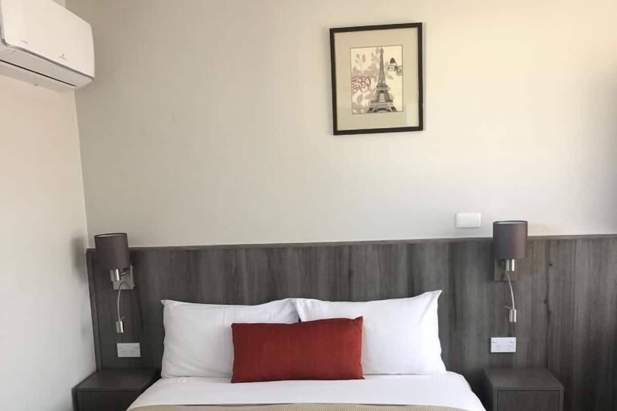 A hotel room with a bed, a framed picture of the Eiffel Tower, two nightstands, and an air conditioning unit.