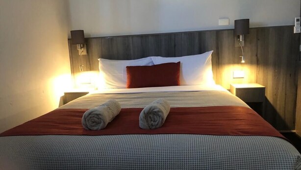 A neatly made bed with two rolled towels, a red pillow, and a wooden headboard. - Bradman Motor Inn (Cootamundra)