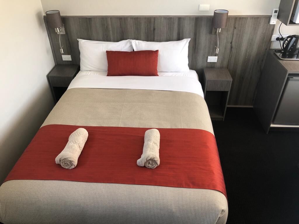 A hotel room with a neatly made bed, a red and white pillow, and two rolled towels on the bed.