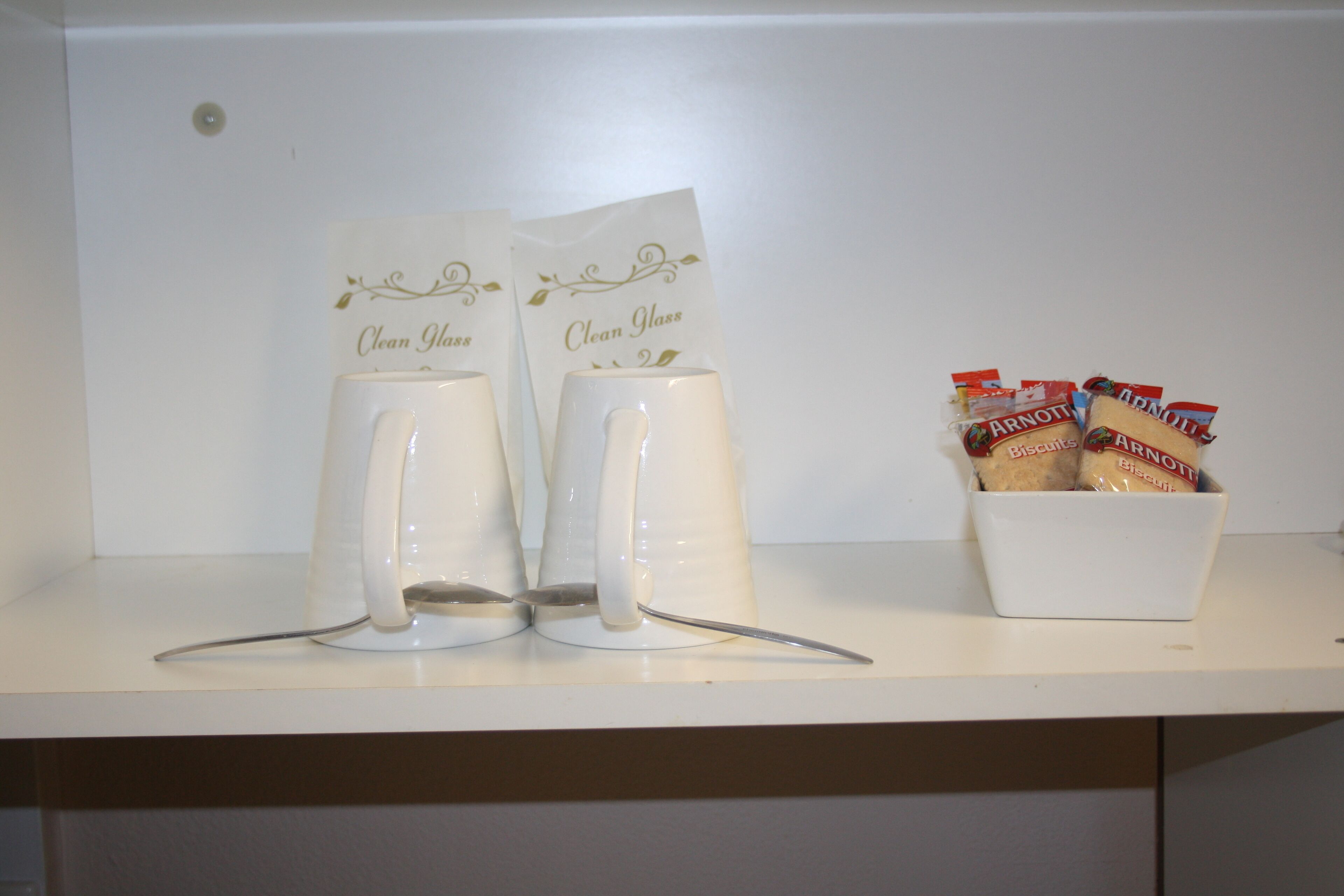 Una estantería blanca con dos tazas blancas etiquetadas “Clean Glass” y un cuenco con paquetes de galletas.