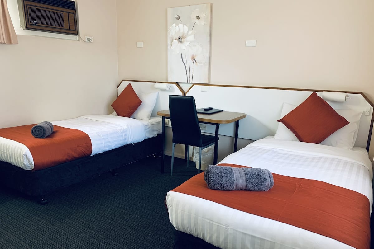 A hotel room with two beds, each with white and orange bedding, a small desk with a chair, and a painting on the wall.