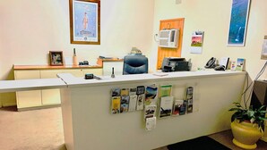 A reception desk with a white counter, a black office chair, a framed picture on the wall, an air conditioning unit, and a potted plant. - Bradman Motor Inn (Cootamundra)