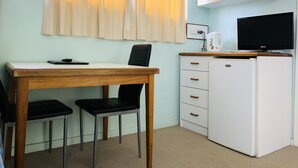 A small, clean room with a wooden desk, two black chairs, a white cabinet with drawers, a small white refrigerator, and a black television on top. - Bradman Motor Inn (Cootamundra)