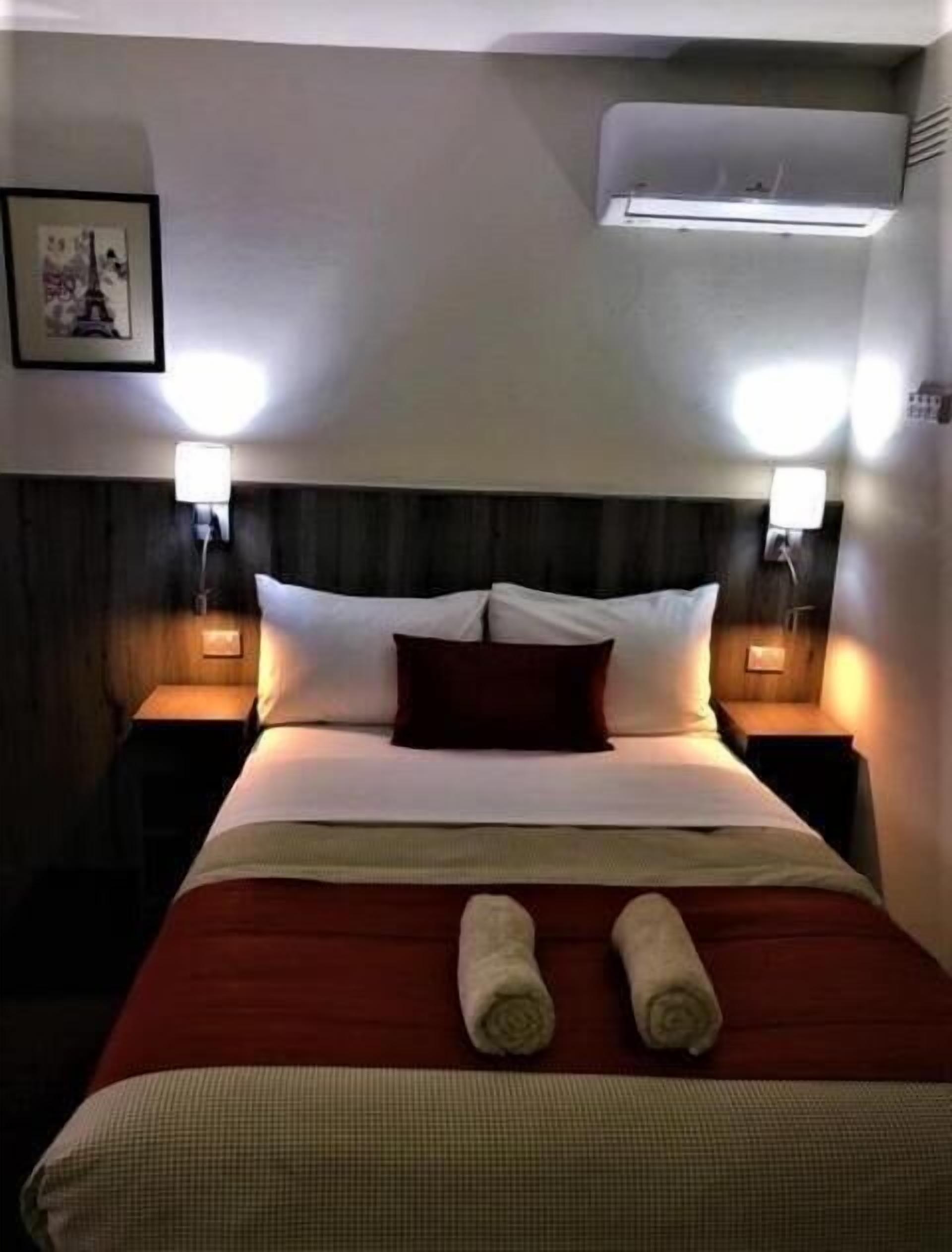 A neatly made bed with two pillows, a red blanket, and two rolled towels. There are two bedside lamps, a framed picture on the wall, and an air conditioning unit above the bed.