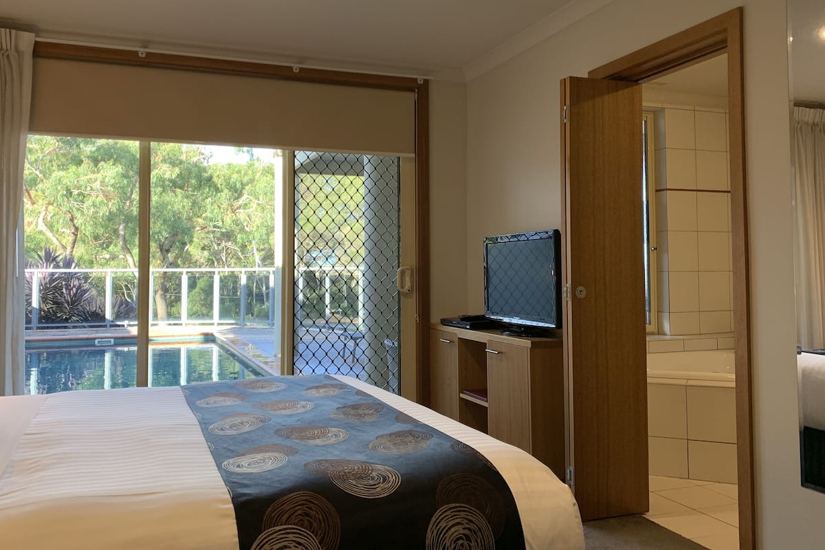 Ein Schlafzimmer mit einem Bett, einem Laptop auf einem Schreibtisch und einer Glastür, die auf einen Balkon mit einem Pool führt.