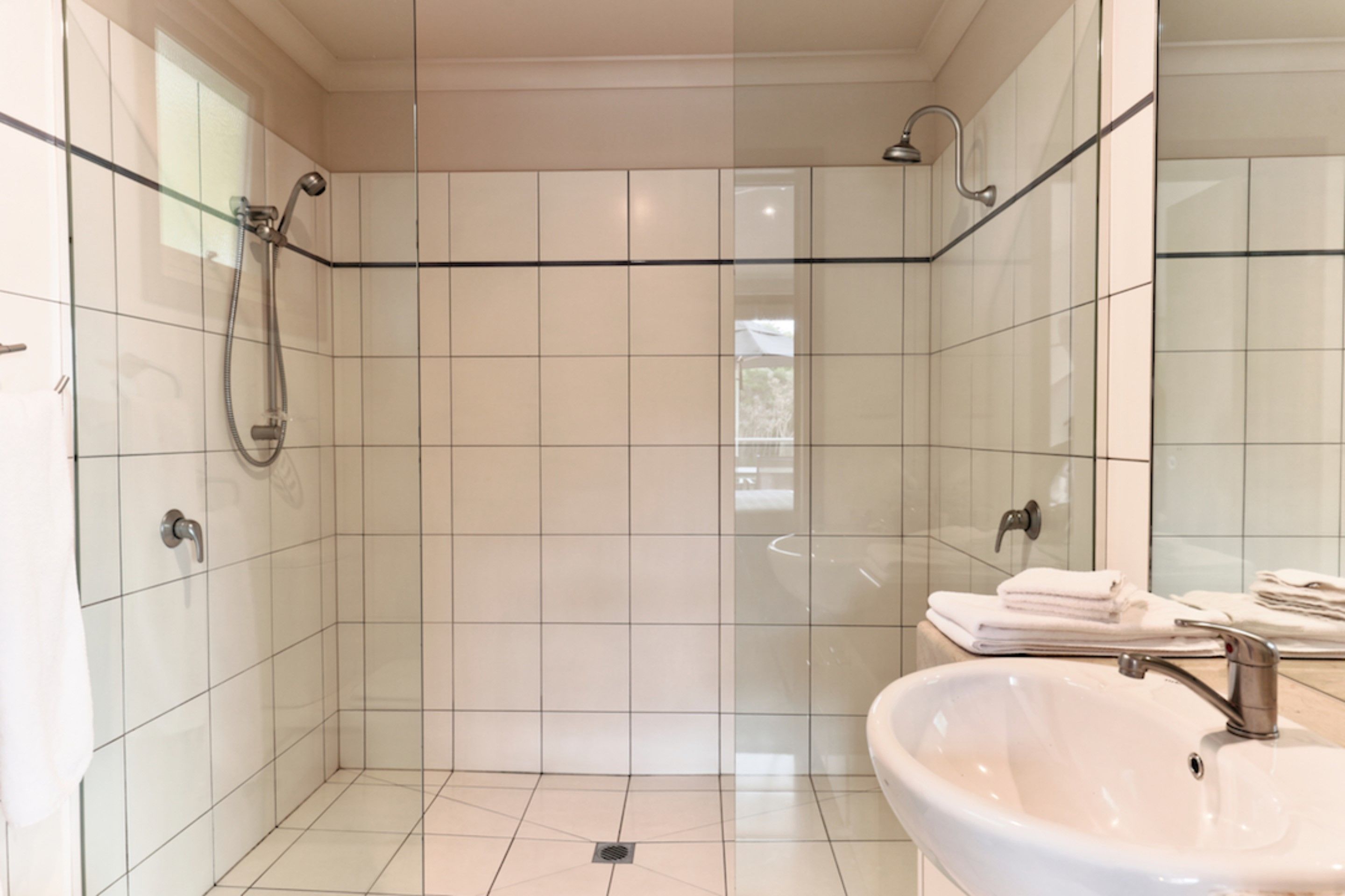 Un baño moderno con paredes de azulejos blancos, cabina de ducha de vidrio, lavabo con espejo y toallas ordenadas.