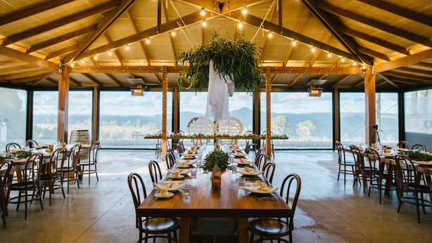 Un amplio comedor con una larga mesa de madera dispuesta para una comida, rodeada de sillas, bajo un alto techo de madera con luces colgantes.