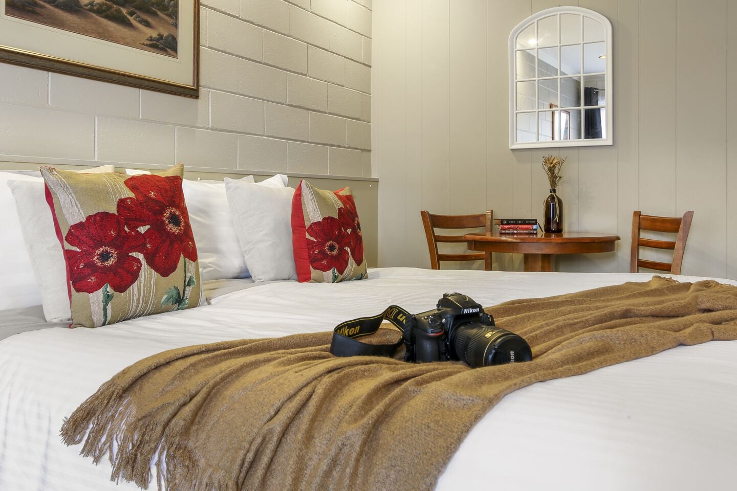 A hotel room with a bed, pillows, a blanket, a camera, a table with books, and a chair.