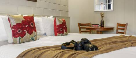 Una habitación de hotel con cama, almohadas, una colcha, una cámara, una mesa con libros y una silla.