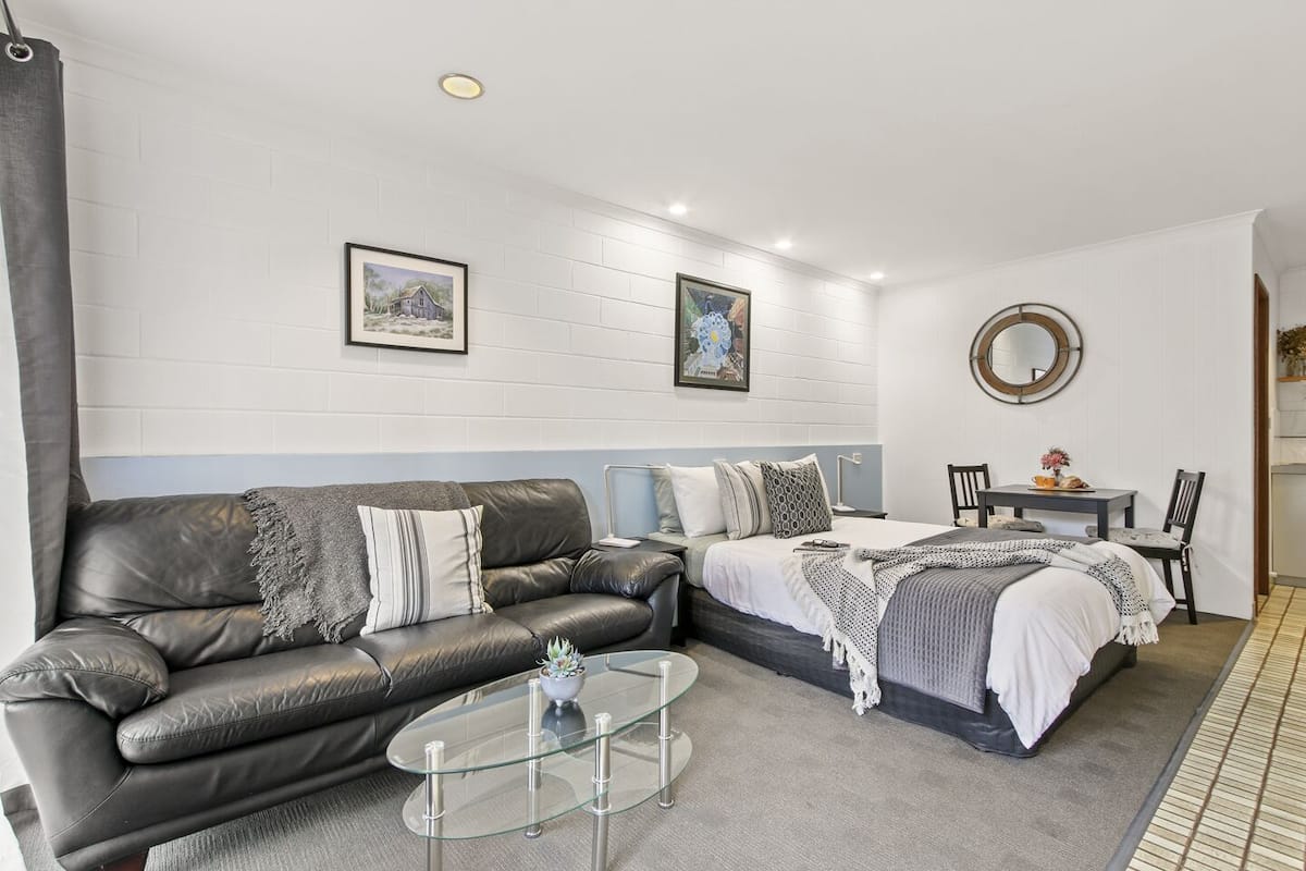 A modern bedroom with a leather sofa, a glass coffee table, a bed with a white headboard, and two framed pictures on the wall.