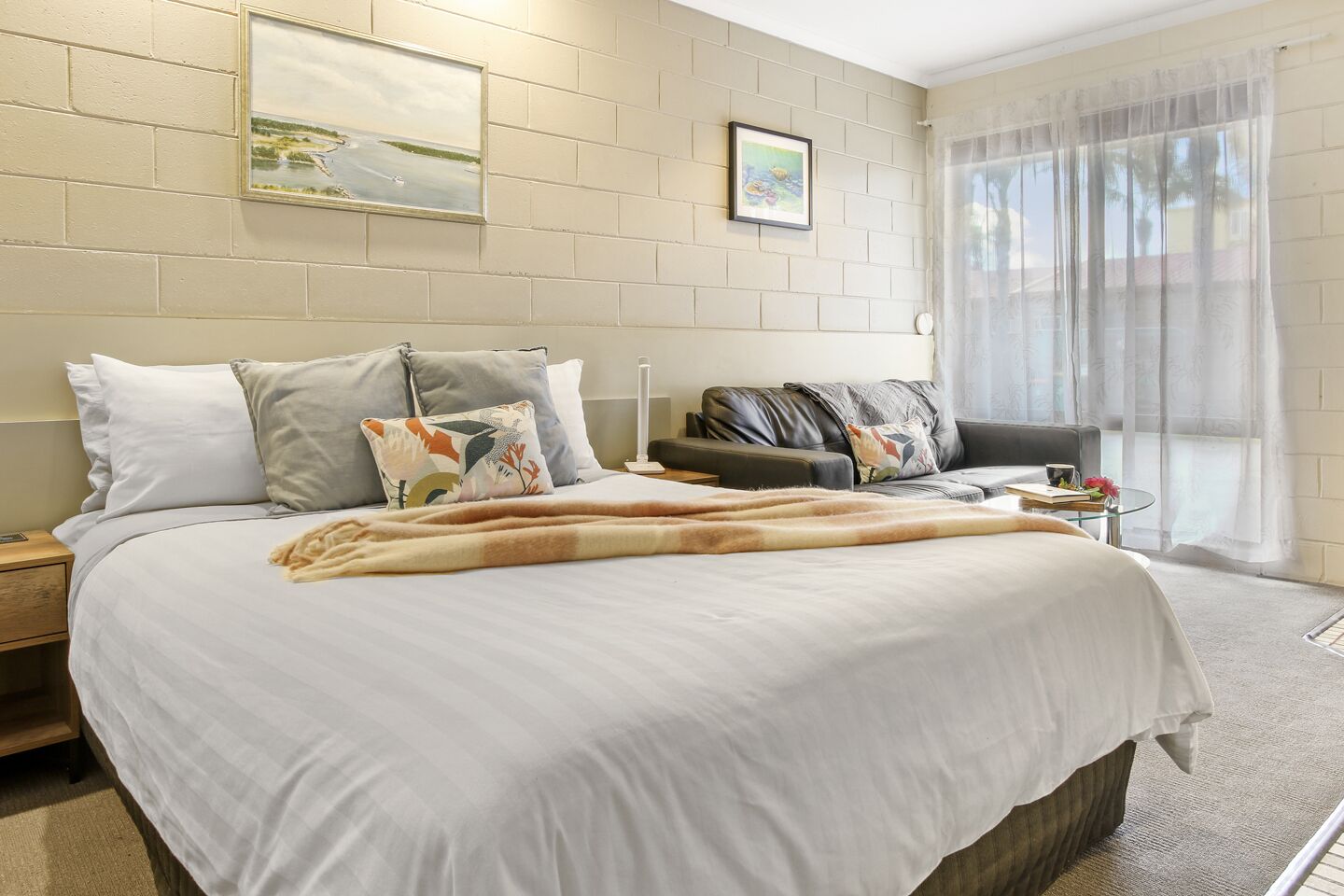A hotel room with a large bed, a sofa, a small table with a lamp, and a window with sheer curtains.