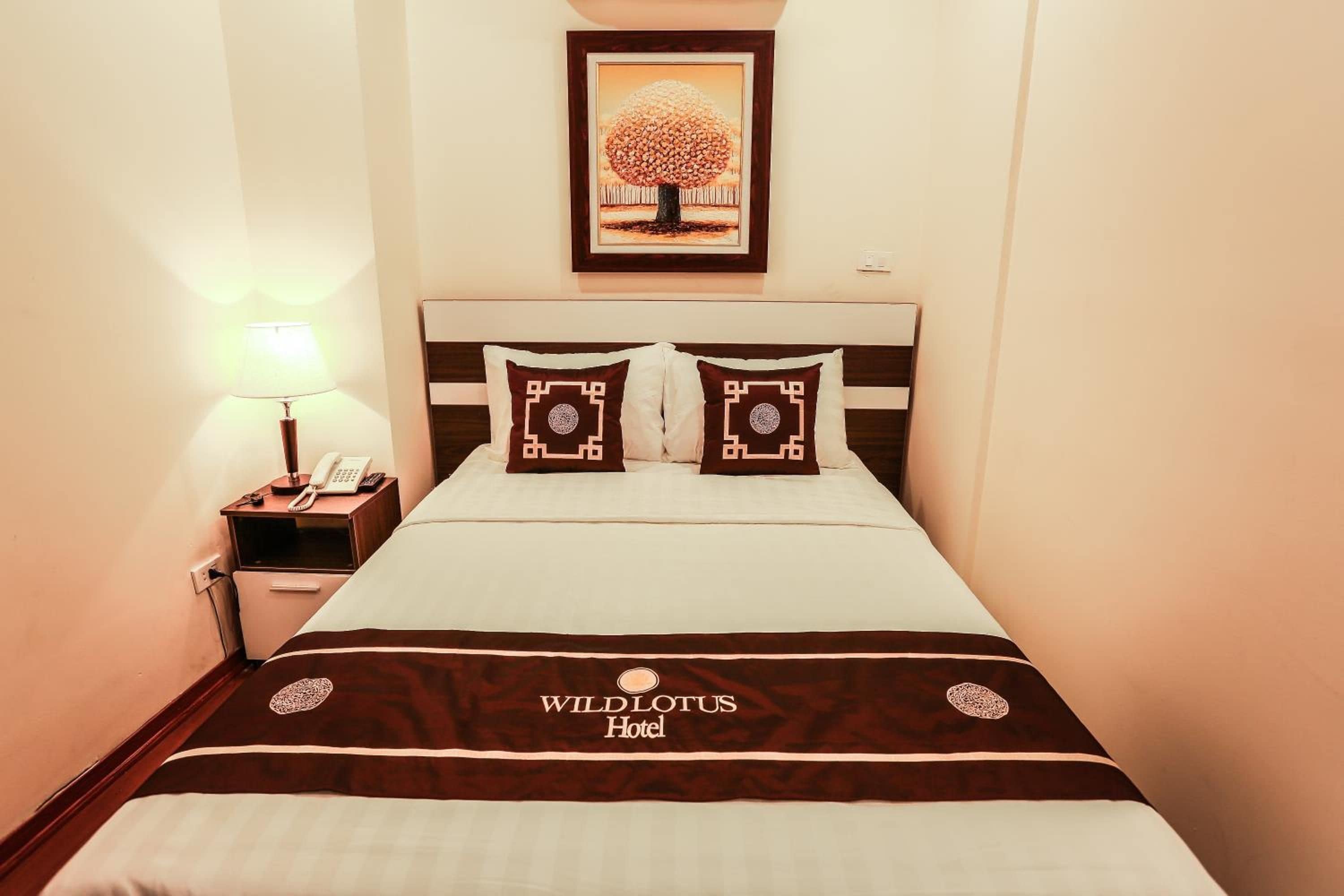 Een hotelkamer met een bed, een nachtkastje met een lamp, een foto in een lijst aan de muur en een sprei met het logo van het hotel.