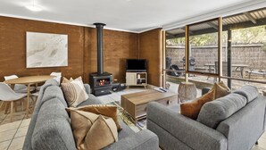 A modern living room with a fireplace, sofa, TV, and large windows overlooking an outdoor area. - Split Point Cottages (Aireys Inlet)