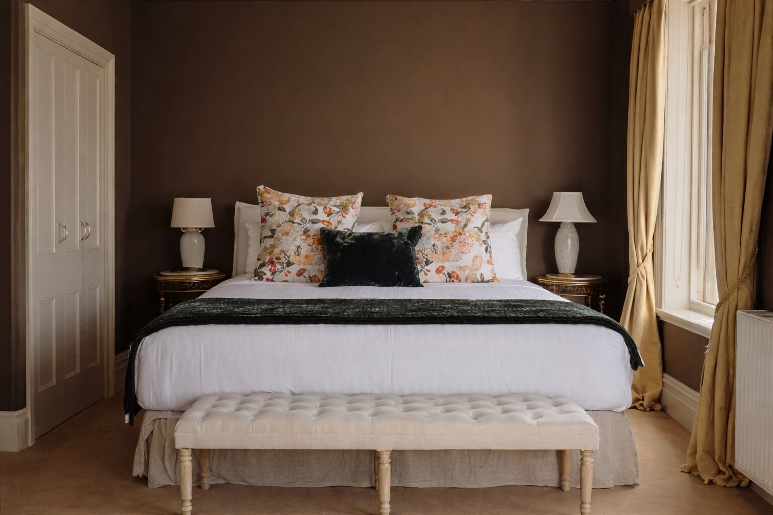 A bedroom with a large bed, two bedside tables, a bench, and a window with curtains.