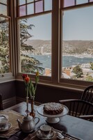 Una mesa de comedor con un pastel, un juego de té y un jarrón con flores, y una vista de una bahía con barcos.