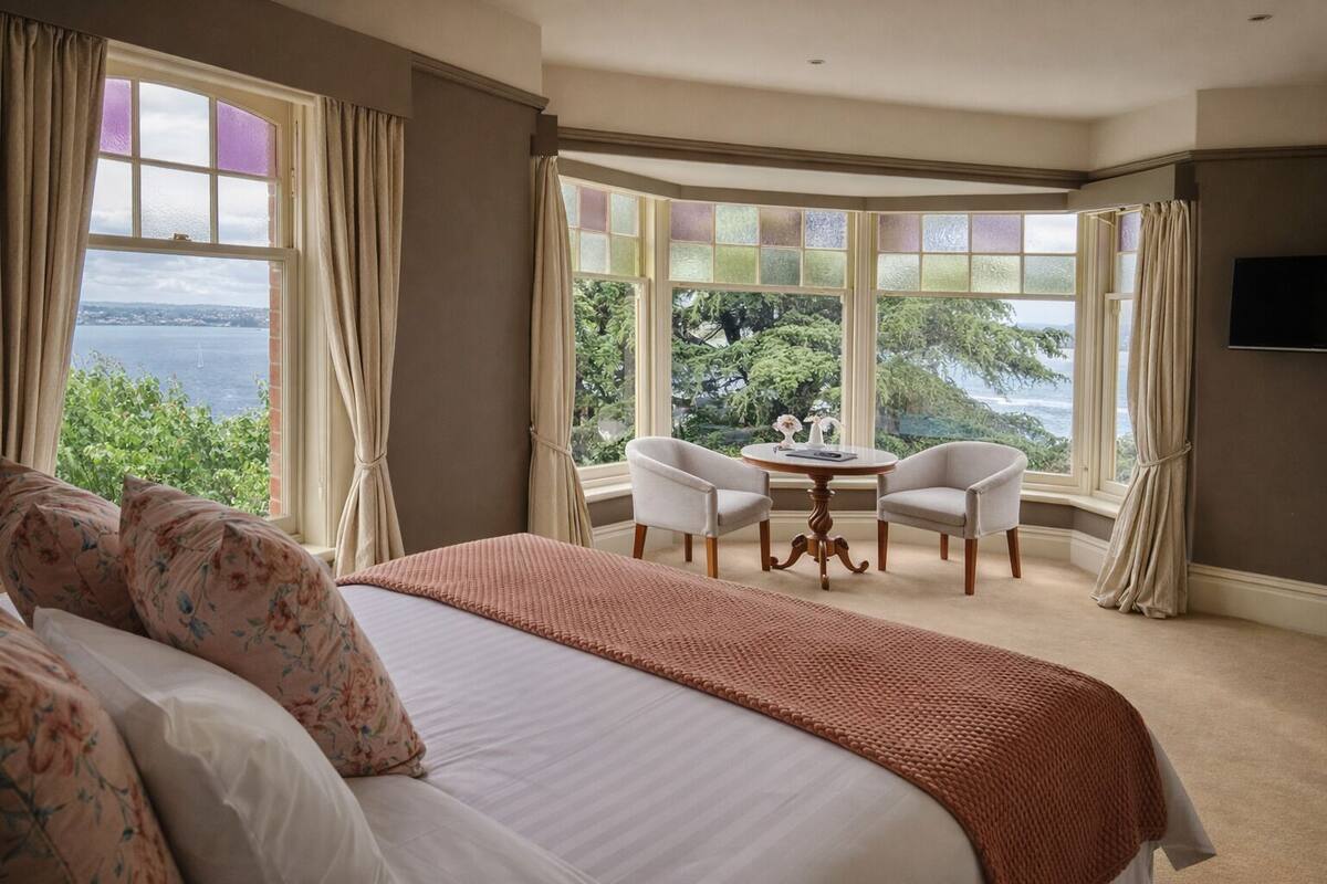 A bedroom with a large bed, two chairs, a small table, and a view of the sea.