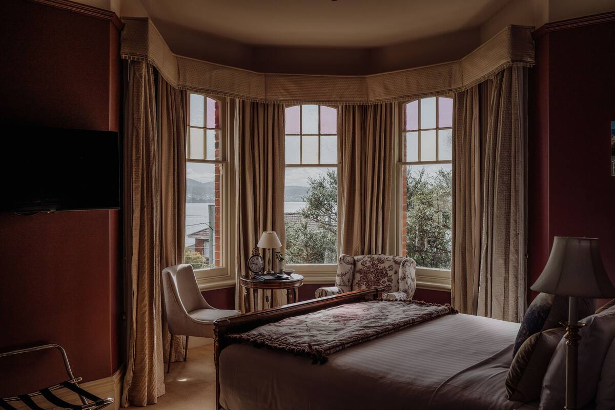 A hotel room with a large bed, a chair, a small table with a lamp, and a TV on the wall.
