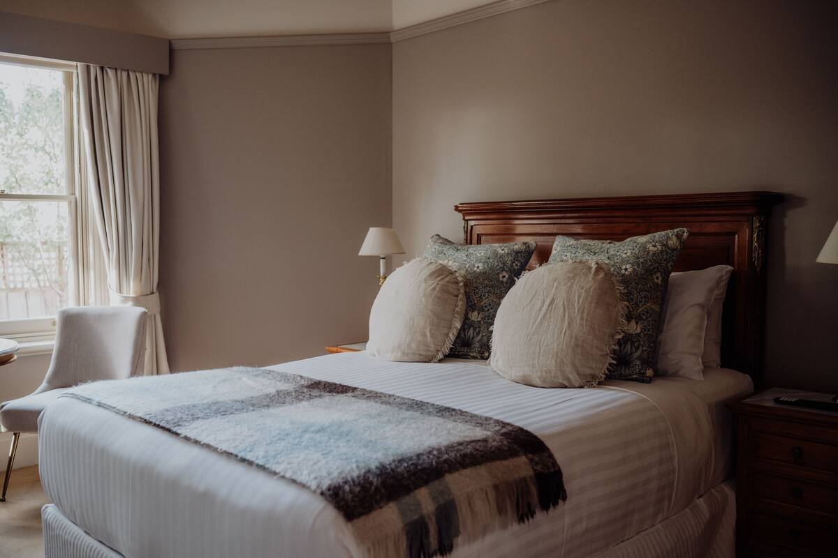 A neatly made bed with a wooden headboard, a bedside table with a lamp, and a chair by the window.