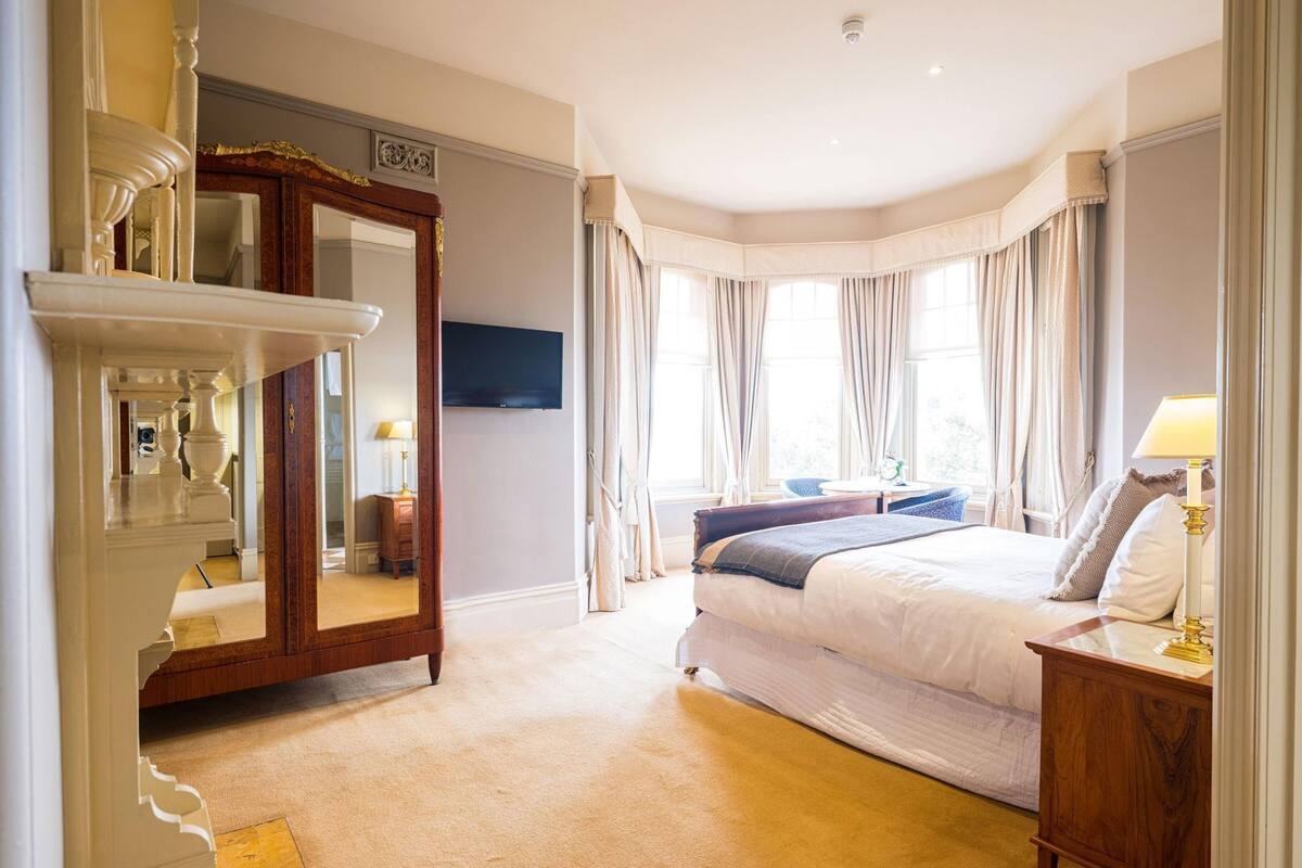 A bedroom with a large bed, a dresser, a mirror, a TV, and a window with curtains.
