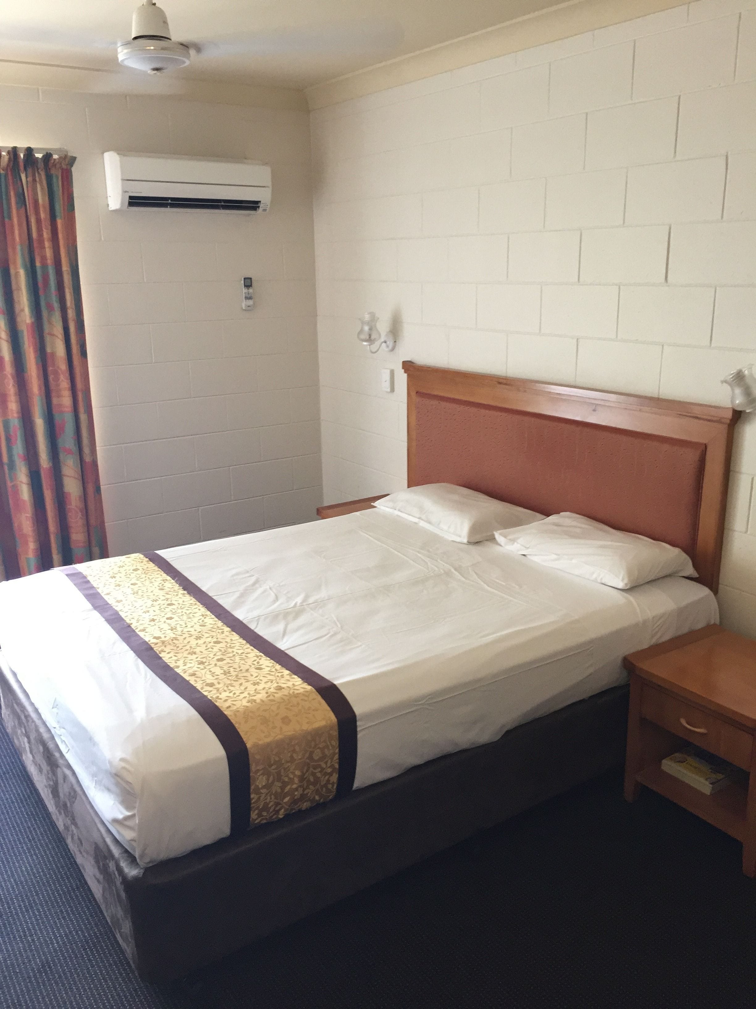 A hotel room with a single bed, a wooden headboard, a nightstand, a bedside table, an air conditioning unit, and a wall-mounted light fixture.