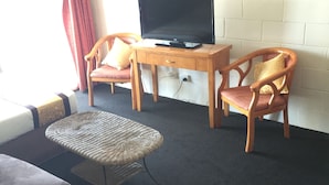 Une pièce comprenant un canapé, deux chaises en bois, une petite table, un téléviseur posé sur un meuble en bois et une fenêtre avec des rideaux rouges.