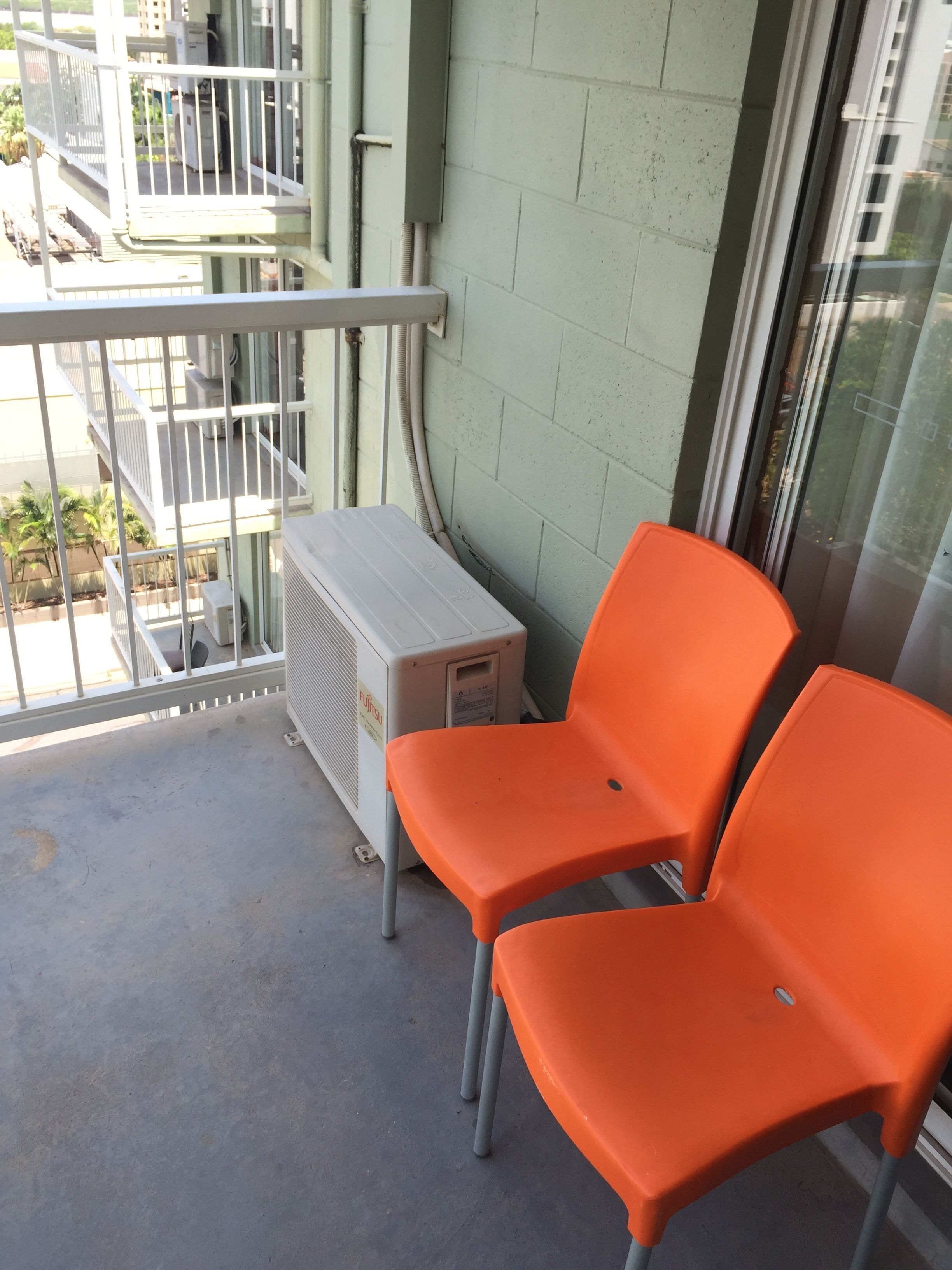 Ein Balkon mit zwei orangefarbenen Stühlen, einer weißen Klimaanlage und Blick auf ein Gebäude mit weißer Brüstung und grüner Wand.