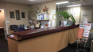 Un comptoir d’accueil en bois, un ordinateur et une plante. Des tableaux encadrés ornent le mur, et une enseigne indique « AATKings ».