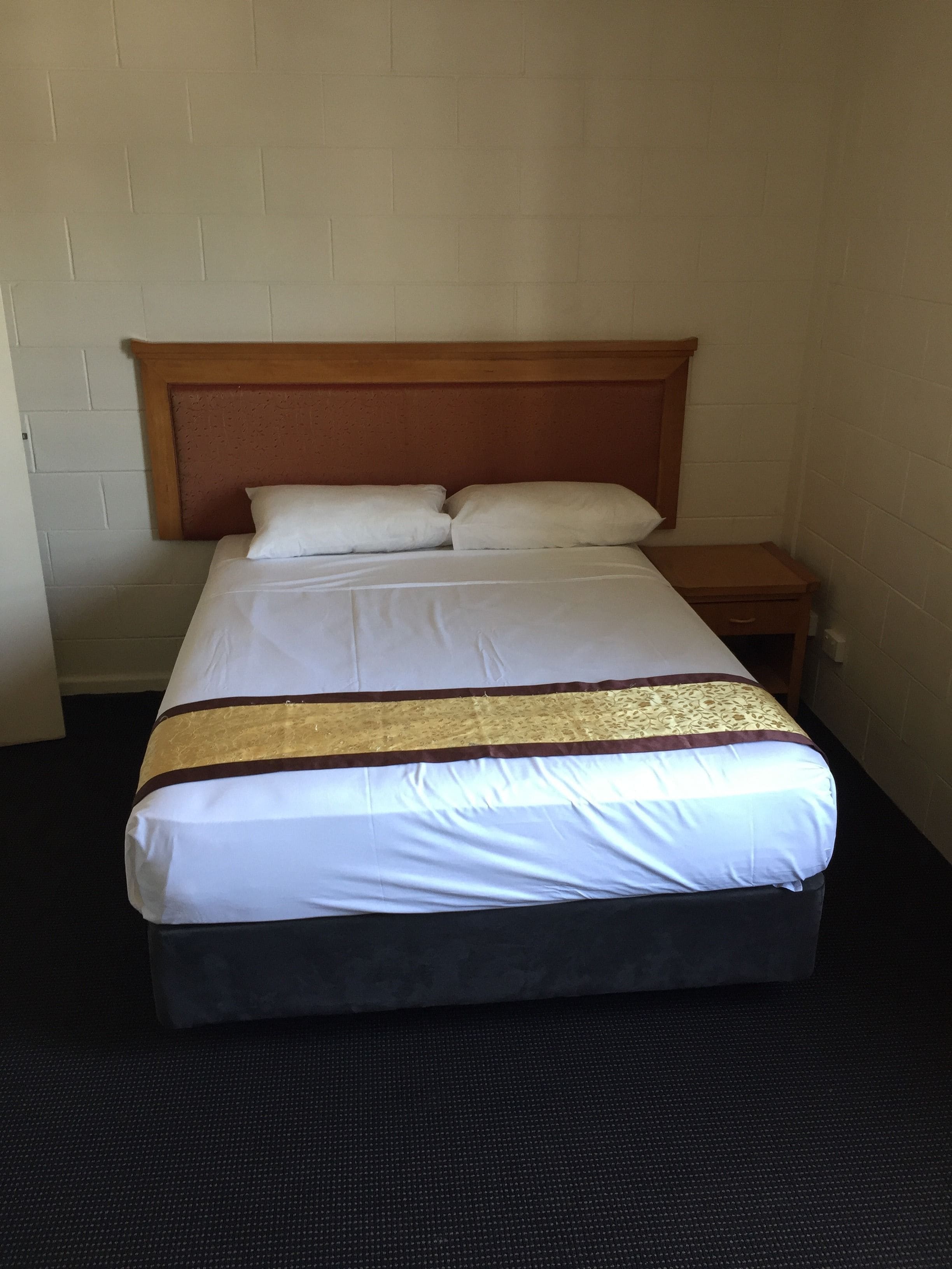 Una camera d'albergo standard con un letto singolo, una testiera in legno, un comodino e un pavimento coperto da moquette scura.