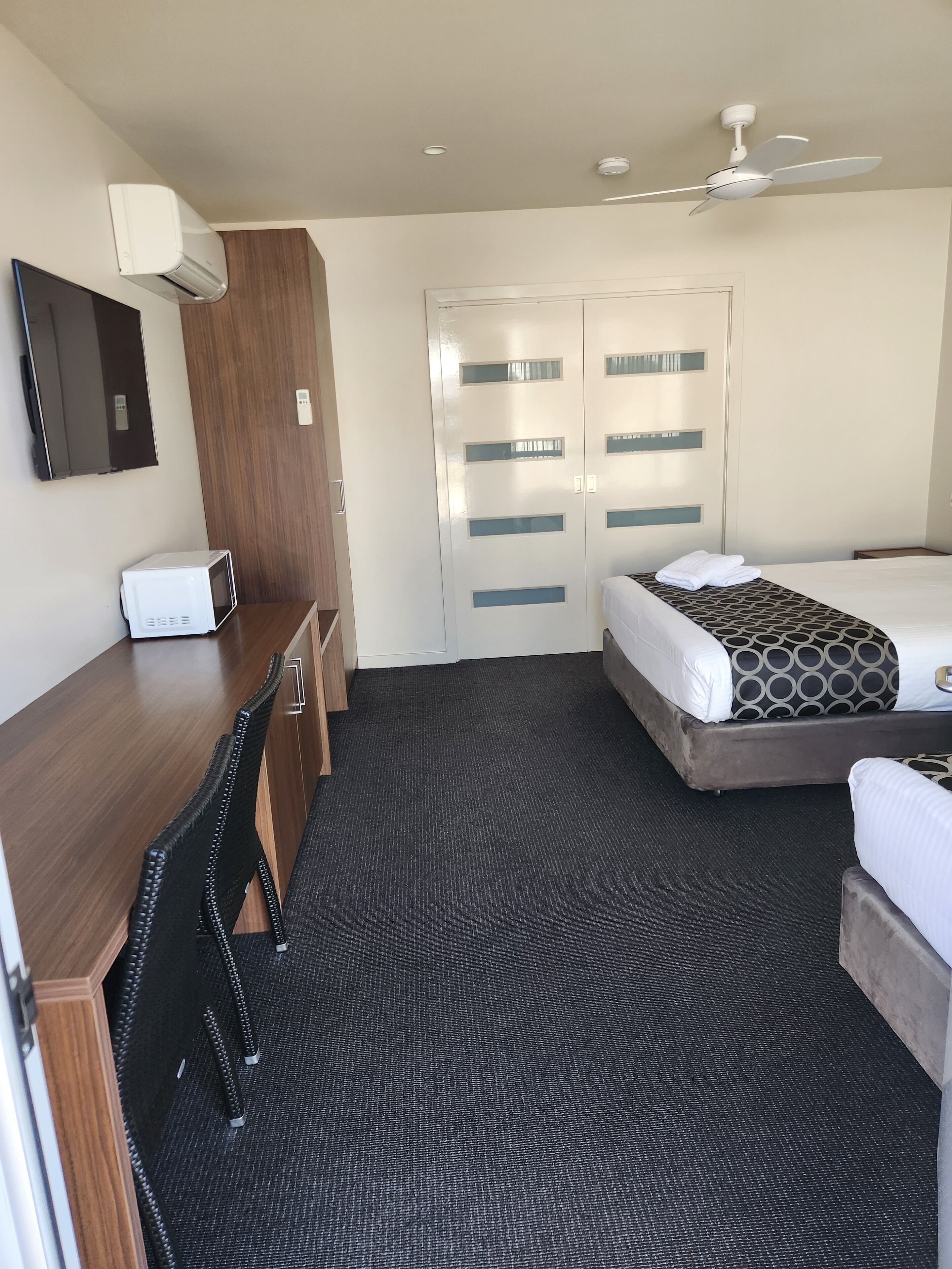 A hotel room with a bed, a desk with a chair, a microwave, a ceiling fan, and a wardrobe.