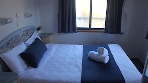 A neatly made bed with a white and dark blue bedspread, a folded towel on the bed, and a window with curtains.