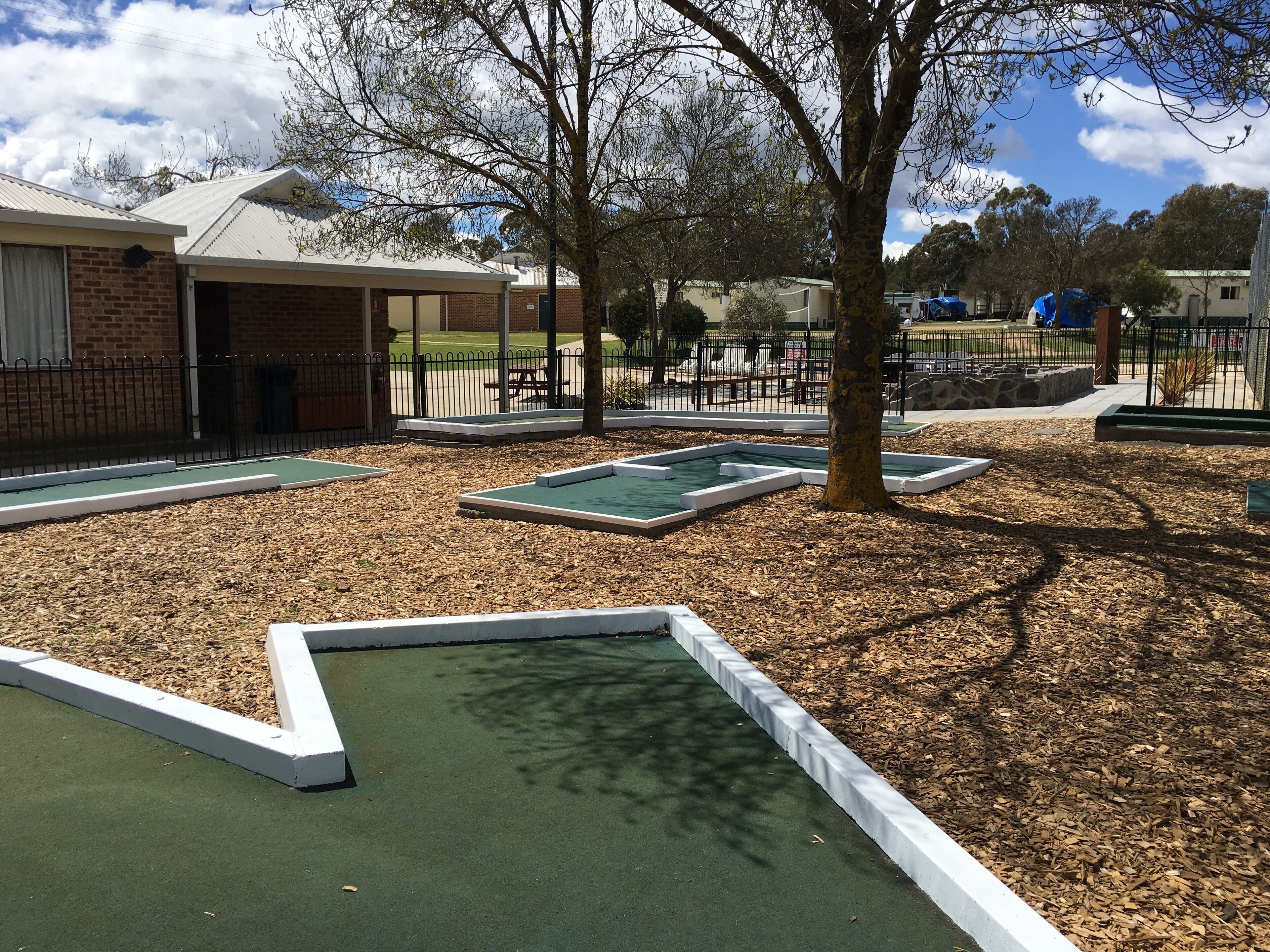 A golf putting area with green turf and white borders, surrounded by trees and a brick building.