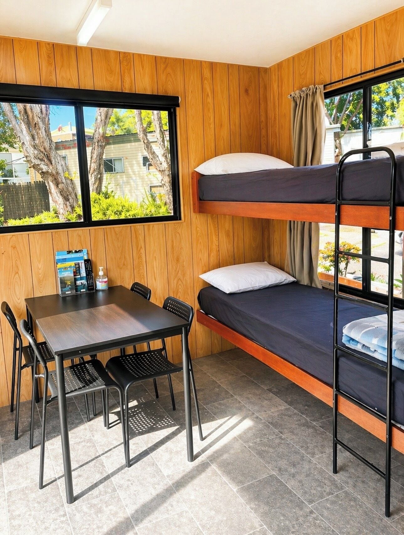 Una cama litera con una mesa y sillas, una ventana con cortinas, y vistas a árboles y edificios.